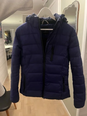 Lila pufferjacka med huva - Snygg lila pufferjacka med huva och dragkedja framtill. Jackan har tre praktiska fickor med dragkedja. Finns en ficka längst med sidan (sista bilden) då den är till för mer ridjacka.  i storlek S! 