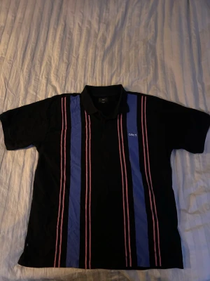 Svart randig skjorta från Obey - Säljer en svart skjorta från Obey med vertikala blå och röda ränder framtill. Skjortan har krage, korta ärmar och en klassisk passform. Perfekt för dig som gillar streetwear och vill sticka ut lite extra.