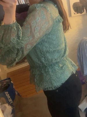 Grön spetsblus med volanger - Snygg grön blus i transparent spets med volangdetaljer och rund halsringning. Blusen har långa ärmar med resår vid ärmslut och en feminin, figurnära passform. Perfekt för dig som gillar romantisk och trendig stil.