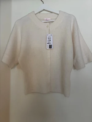 Krämvit kortärmad kofta  - Söt krämvit kofta från Gina med korta ärmar och rund halsringning. Koftan har ribbade detaljer vid ärmslut och nederkant samt knäppning framtill med fem knappar. Perfekt att styla över en topp eller klänning. Passar som en xs/s