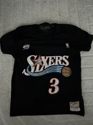 Svart Sixers Iverson baskettröja - Säljer en svart baskettröja från Mitchell & Ness med stort Sixers-tryck och nummer 3 på framsidan samt Iverson och nummer 3 på ryggen. Tröjan har NBA-logga och 2001-detalj, samt klassisk meshstruktur för bra ventilation. Perfekt för basketfans!