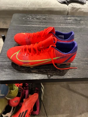 Nike Mercurial röda fotbollsskor - Säljer ett par Nike Mercurial fotbollsskor i en riktigt snygg röd färg med neonfärgade detaljer och blå insida. Skorna har snörning, låg profil och är tillverkade i syntetmaterial för bästa grepp och känsla på planen. Perfekta för dig som vill sticka ut på fotbollsplanen. Ej elites.