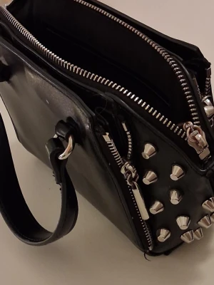 Svart handväska med nitar och dragkedjor - Cool svart handväska i glansigt material med silverfärgade nitar och detaljer. Väskan har flera dragkedjor. Är från zara PRIS kan diskuteras 💕