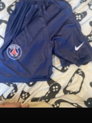 Blåa PSG shorts från Nike - Säljer ett par mörkblå PSG shorts från Nike med klubbens logga på ena benet och Nike swoosh på det andra. Shortsen har resår i midjan och är gjorda i ett lätt och sportigt material, perfekta för träning eller match.