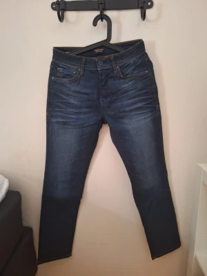Jack & Jones jeans 27/30 - Snygga mörkblå slim fit jeans med klassisk femficksdesign och orangea sömmar. Jeansen har en lätt tvättad look och är perfekta för en trendig vardagsstil. Tillverkade i stretchigt denim för extra komfort.