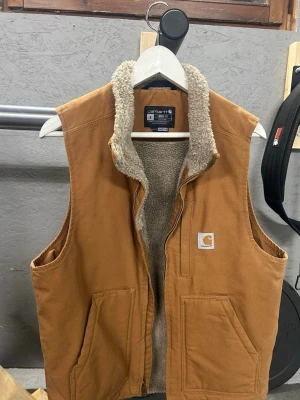 Brun Carhartt dunväst med teddyfoder - Snygg brun dunväst från Carhartt med mjukt teddyfoder på insidan och kragen. Västen har två stora fickor framtill och dragkedja. Perfekt för lager-på-lager och ger en avslappnad streetwear-look. Klassisk Carhartt-logga på bröstet.