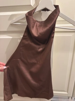 Brun halterneck topp i satin - Snygg brun halterneck topp i glansigt satinmaterial. Toppen har hög krage och är ärmlös med en smickrande passform. Perfekt för dig som vill ha en trendig och stilren look.