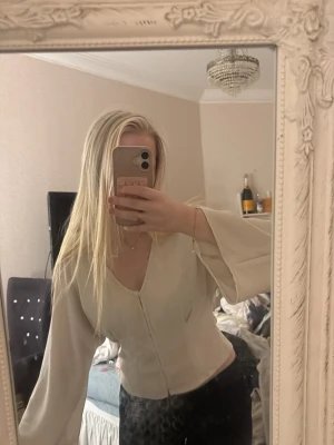 Krämvit blus med vida ärmar - Snygg krämvit blus med v-ringning och knappar framtill. Blusen har långa, vida ärmar och är figursydd i midjan . Hör av vid frågor! 