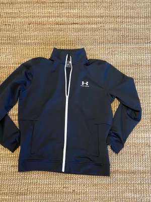 Svart Under Armour Zip-tröja - Säljer en stilren svart zip-tröja från Under Armour med vita detaljer. Tröjan är i mycket fint skick och har endast använts som vardagsplagg vid ett fåtal tillfällen. Nypris: 700kr