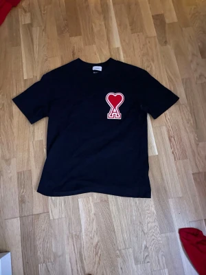 Svart Ami Paris t-shirt med hjärtdetalj - Svart t-shirt från Ami Paris med en stor röd hjärtformad Ami-logga på bröstet. Klassisk rak passform och rund halsringning. Tillverkad i mjuk bomull som känns skön mot huden. Perfekt för dig som gillar stilrena plagg med en twist.
