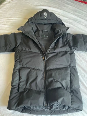 Svart pufferjacka från 8848 Altitude - Snygg svart pufferjacka från 8848 Altitude med quiltad design och huva. Jackan har dragkedja framtill och logga på huvan. Perfekt för kalla dagar och har en modern, oversized look.