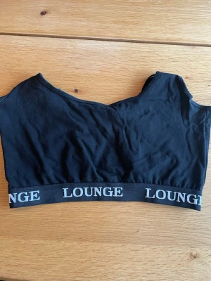 Svart Lounge crop top med korsade band - Svart crop top från Lounge med bred resår och vit logotyp runt om. Toppen har tunna, justerbara axelband som korsas i ryggen för en snygg detalj. Tillverkad i mjukt och stretchigt material som passar perfekt för chill dagar hemma.