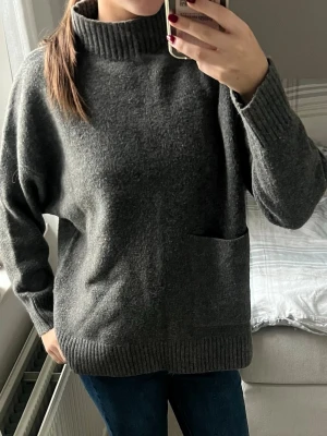 Mörkgrå stickad polotröja - Mysig mörkgrå stickad tröja med hög ribbad polokrage och en stor ficka framtill. Tröjan har avslappnad passform, långa ärmar och breda ribbade muddar vid ärmslut och nederkant. Perfekt för kalla dagar och enkel att matcha med jeans.