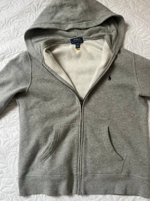 Grå hoodie från Polo Ralph Lauren - Snygg grå hoodie från Polo Ralph Lauren med dragkedja och två fickor framtill. Insidan är fodrad med mjuk vit fleece och den har en klassisk huva. Liten broderad logga på bröstet. Perfekt för en chill och avslappnad stil.