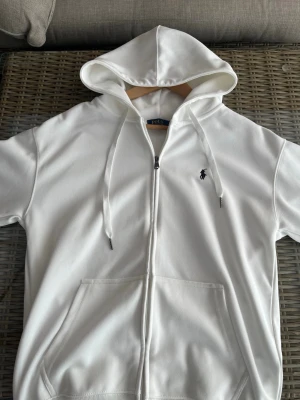Vit hoodie från Polo Ralph Lauren - Säljer en clean vit hoodie från Polo Ralph Lauren med dragkedja, huva med dragsko och kängurufickor. Klassisk liten logga broderad på bröstet. Perfekt till chill vardagsstil och enkel att matcha med allt. Materialet är mjukt och skönt bomullstyg. Jag är 183 och väger 75 kg och den passar som en hoodie ska vara 
