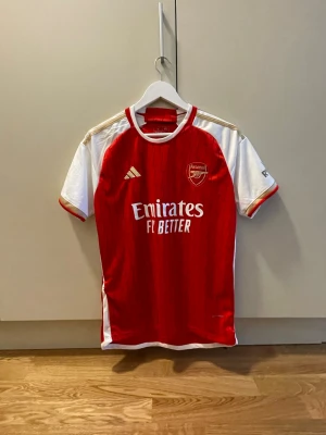 Arsenal matchtröja Adidas röd/vit - Snygg Arsenal matchtröja från Adidas i rött och vitt med gulddetaljer. Tröjan har klubbmärke och sponsortryck på bröstet, samt klassiska Adidas-ränder på axlarna. Tillverkad i lätt och ventilerande material, perfekt för fotboll eller träning.
