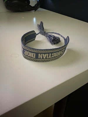 Blått Christian Dior tygarmband - Snyggt blått tygarmband från Christian Dior med broderad logga i vitt. Armbandet har justerbar knytning med tofsar i ändarna och en cool, bohemisk känsla. Perfekt accessoar för att lyfta din outfit med en exklusiv touch.