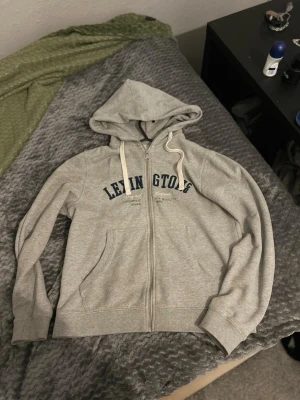 Grå zip hoodie från Lexington - Snygg grå zip hoodie från Lexington med mörkblå text på bröstet och vita dragsnören i huvan. Klassisk modell med fickor framtill och mjukt material, perfekt för chill dagar. Dragkedja hela vägen och ribbade muddar vid ärmar och nederkant.