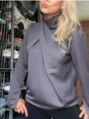 Mörkgrå asymmetrisk blus - Snygg mörkgrå blus med unik asymmetrisk krage och omlott-detalj framtill. Tillverkad i ett slätt, glansigt material som ger en elegant känsla. Långärmad modell som passar perfekt till både jeans och kjol. Märke ej synligt.