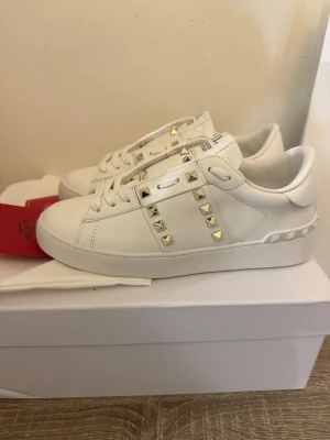 Vita Valentino Garavani sneakers - Snygga vita sneakers från Valentino Garavani med ikoniska guldfärgade nitar längs sidorna och på plösen. Skorna är i läder med klassisk låg siluett och vita skosnören. Perfekta för dig som vill sticka ut med en exklusiv touch.