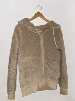 Beige teddyhoodie med dragkedja - Mysig beige hoodie från Monki i teddyfleece med dragkedja framtill, huva med dragsko och två fickor. Muddar vid ärmslut och nederkant ger en skön passform. Perfekt för en avslappnad och trendig look.