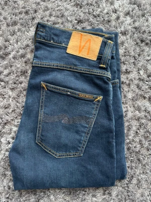 Nudie jeans grim Tim - Snygga blå jeans från Nudie Jeans med klassisk femficksdesign och kontrastsömmar. Jeansen har en rak passform och är tillverkade i mjuk bomullsdenim. Bakfickan har Nudie Jeans ikoniska broderade vågmönster och läderpatch i midjan.