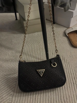 Svart mini axelväska - Svart mini-axelväska i strukturerat material med präglat mönster. Guldfärgade detaljer och tunn kedjerem ger en nätt men markerad look. Dragkedjestängning upptill och kompakt storlek – perfekt för mobil, kort och nycklar.❤️