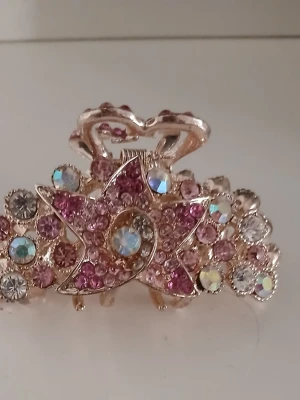 Hårklämma med rosa och vita stenar - Superfin hårklämma i guldmetall dekorerad med glittrande rosa och vita strasstenar i blommönster. Klämman har en elegant form med hjärtformade detaljer och passar perfekt för att ge frisyren extra bling. Materialet är metall och strass.
