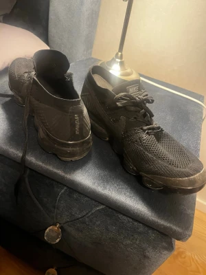 Nike Air Vapormax svarta sneakers - Svarta Nike Air Vapormax sneakers med stickad ovandel i mesh och synlig dämpande sula. Skorna har snörning och diskret Nike-logga på sidan. Snygg och sportig design som passar till streetwear och vardagsstil.