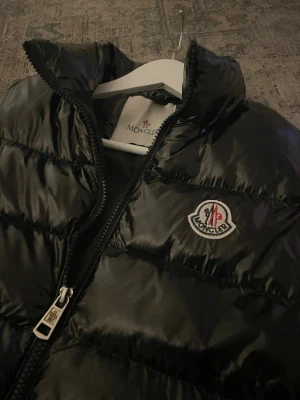 Svart dunväst från Moncler - Snygg svart dunväst från Moncler med glansig finish och hög krage. Västen har dragkedja framtill, två fickor med dragkedja och Moncler-logga på bröstet. Perfekt för lager-på-lager och streetstyle.