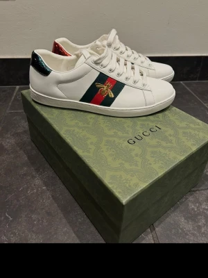 Gucci Ace vita sneakers med bi - Vita Gucci Ace sneakers i läder med klassiska gröna och röda ränder på sidan samt broderad guldfärgad bi. Skorna har metallic-detaljer i rött och grönt på hälen och vita snören. Perfekta för dig som vill sticka ut med lyxig streetstyle.