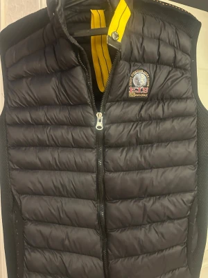 Svart dunväst från Parajumpers - Snygg svart dunväst från Parajumpers med gul detalj vid kragen och broderad logotyp på bröstet. Västen har quiltad design, dragkedja framtill och är lätt vadderad för extra värme. Perfekt att slänga över en hoodie eller tröja för en cool look.