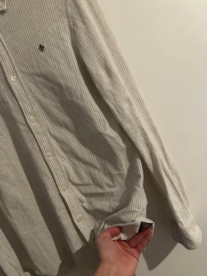 Randig beige skjorta från Morris - Säljer en stilren beige skjorta från Morris med tunna vita ränder och klassisk krage. Skjortan har lång ärm, knappar framtill och en liten broderad logga på bröstet. Nypris 1699 och mitt pris 249 ✨, Tillverkad i mjuk bomull som känns skön mot huden.