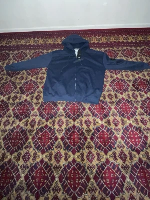 Mörkblå zip hoodie med huva - En enkel och stilren mörkblå hoodie med dragkedja framtill och huva med dragsko. Hoodien har långa ärmar och ribbade muddar vid ärmslut och nederkant. Perfekt för en avslappnad look och passar till det mesta. Materialet ser ut att vara mjukt och bekvämt, troligen bomullsblandning.