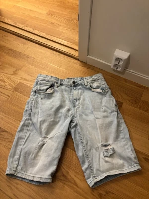 Ljusblå jeansshorts med slitningar - Säljer ett par ljusblå jeansshorts med slitna detaljer på framsidan. Klassisk femficksmodell med knapp och dragkedja. Perfekta för sommaren och ger en avslappnad streetstil.