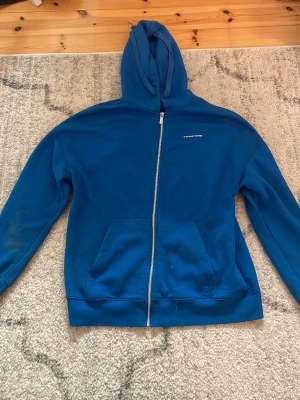 Blå zip hoodie  - Blå hoodie med dragkedja. Klassisk modell med huva och två stora fickor framtill. Liten vit logga på bröstet. Perfekt för chill dagar eller när du vill ha en enkel och snygg look. Mjuk bomullskänsla och relaxed fit.