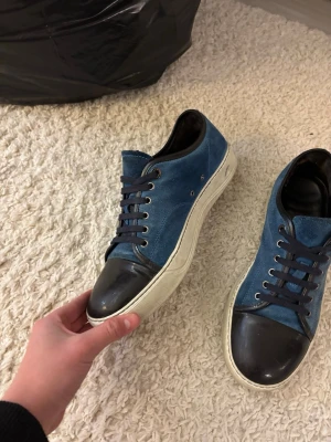 Blå sneakers från Lanvin med svart tå - Snygga blå sneakers från Lanvin med svart tåparti i läder och vita sulor. Skorna har mörkblå skosnören och silverfärgade öljetter. Klassisk låg modell med en stilren design som passar till många outfits.