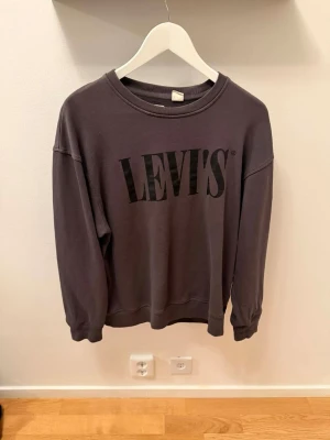 Mörkgrå sweatshirt från Levi's - En stilren mörkgrå sweatshirt från Levi's med stor svart logga framtill. Tröjan har rund halsringning, långa ärmar och ribbade muddar vid ärmslut och nederkant. Perfekt för en avslappnad och trendig look.
