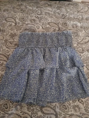 Blommig blå volangkjol - Supersöt kort kjol med blått och vitt blommönster och bred smock i midjan. Kjolen har flera volanger som ger en luftig och lekfull känsla. Perfekt för dig som gillar en feminin och avslappnad stil. Kan skicka fler bilder vid förfrågan 