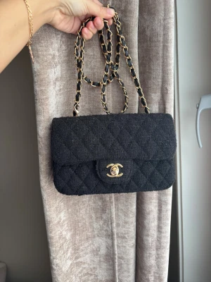 Svart Chanel quiltad handväska - Snygg svart handväska från Chanel i klassisk quiltad design med guldiga detaljer och kedjerem med insytt läder. Väskan är tillverkad i tyg och har den ikoniska CC-loggan framtill. Perfekt för dig som vill ha en tidlös och lyxig accessoar.