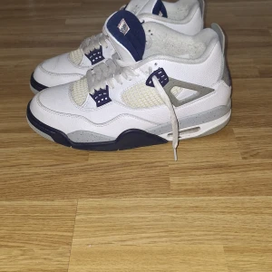 Nike Air Jordan 4 Retro Vit/Marinblå - Säljer ett par Nike Air Jordan 4 Retro i vitt och marinblått. Skorna har detaljer i grått med stänk, meshpaneler och klassisk Jumpman-logga på hälen. Ovandelen är i skinn och de har snörning samt en chunky sula för extra streetstyle.