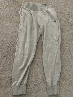 Grå mjukisbyxor från Puma - Snygga grå mjukisbyxor från Puma med resår i midjan och dragsko. Byxorna har ribbade muddar vid bensluten och två sidofickor. Tillverkade i mjukt och bekvämt material, perfekta för chill eller träning. Puma-logga broderad på vänster ben. Den har en liten ljus fläck (se bild 4)