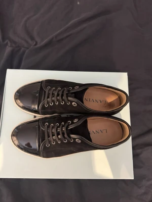 Svarta sneakers från Lanvin - Snygga svarta sneakers från Lanvin med mocka på ovandelen och glansig tå i lack. Skorna har vit sula, svarta skosnören och metallfärgade öljetter. Klassisk låg modell som passar till många olika stilar. Strl är 43 men passar dig som har 44, Ny prisa 5000, mitt pris 3500 men priset kan diskuteras vid snabb affär, hör av vid frågor 
