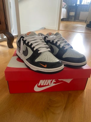 Nike Dunk Low svart/vit med orange detalj - Nike Dunk Low sneakers i svart och vitt läder med vit sula och snörning. Skorna har en orange mini-swoosh på tån och klassisk Nike-logga på hälen. Perfekt för dig som gillar stilrena och sportiga sneakers med en twist.