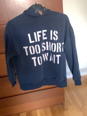 Mörkblå hoodie med texttryck - Säljer en mörkblå hoodie med vit text på framsidan där det står 'LIFE IS TOO SHORT TO WAIT'. Hoodien har huva, ribbade muddar och är gjord i mjukt sweatshirtmaterial. Perfekt för en avslappnad och cool stil.