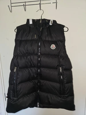 Svart dunväst från Moncler med huva - Stilren svart dunväst från Moncler med huva och dragkedja framtill. Västen har två praktiska fickor med dragkedja och en vit detalj på huvan. Klassisk Moncler-logga på bröstet och justerbar dragsko vid halsen. Perfekt för lager-på-lager och streetstyle.