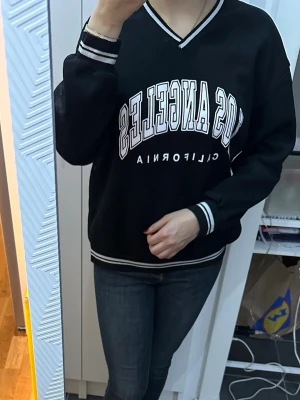 Svart sweatshirt med Los Angeles-tryck - Cool svart sweatshirt med vitt Los Angeles-tryck och texten California. Tröjan har V-ringning och vita ränder på muddar och nederkant. Oversized passform och sportig collegekänsla, perfekt till jeans eller leggings.