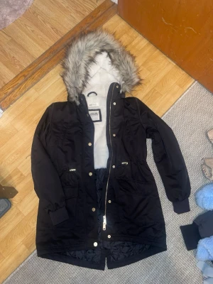 Svart parkasjacka från H&M med fuskpäls - Snygg svart parkasjacka från H&M med mjukt vitt foder och huva med fuskpäls. Jackan har dragkedja och tryckknappar i guld, två stora fickor framtill och ribbade muddar vid ärmsluten. Perfekt för kalla dagar och riktigt mysig att bära.