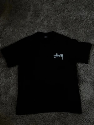 Svart Stüssy t-shirt med logga - Svart t-shirt från Stüssy med klassisk vit logga på bröstet. T-shirten har rund hals och korta ärmar, perfekt för dig som gillar streetwear och enkel stil. Materialet är mjuk bomull som känns skönt mot huden.
