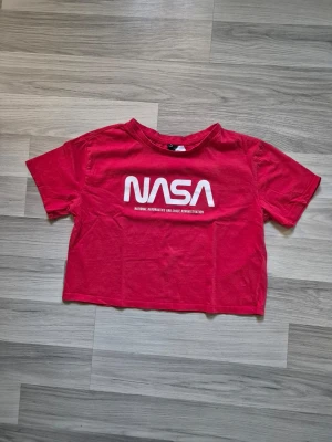 Röd croppad NASA t-shirt - Cool röd t-shirt med vit NASA-logga på bröstet. Modellen är croppad och har korta ärmar. Perfekt för dig som gillar space vibes och vill ha en avslappnad look. Materialet är mjuk bomull som känns skönt mot huden.